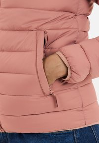 Puffet pink jakke med vertikale quiltede sektioner, udstyret med en lynlås-lomme og en tætsiddende manchet. Glat tekstur, letvægtsmateriale.