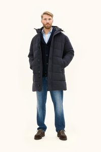 Czarna kurtka puchowa z kapturem, zamkiem błyskawicznym i zapięciem na guziki, o pikowanym wzorze. Zestawiona z granatowym swetrem, jasnoniebieską koszulą i dżinsami.