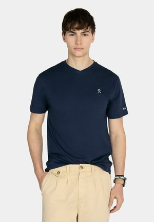 Jeune homme portant un t-shirt bleu marine à col en V avec un petit logo sur la poitrine et un pantalon beige, debout avec une main dans la poche.