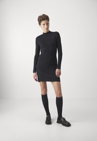 Rochie tricotată neagră cu model de nervuri, cu mâneci lungi și guler înalt, având o siluetă ajustată și o margine deasupra genunchiului. Ciorapi negri până la genunchi și pantofi.