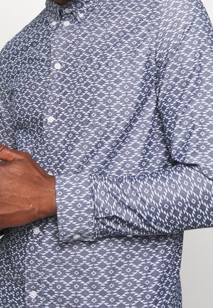 Man wearing blue patterned long-sleeve button-up shirt with white geometric shapes, showing torso and folded hands.