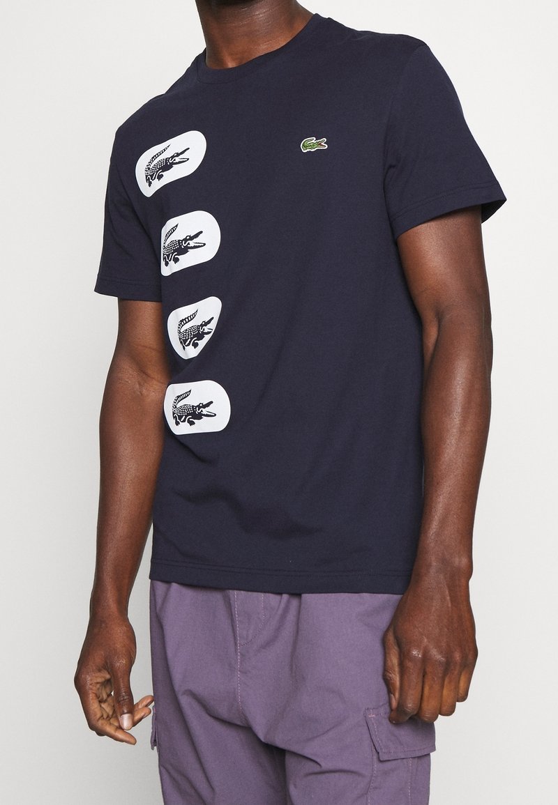 Homme portant une chemise bleu marine à manches courtes avec quatre écussons blancs avec des logos de crocodile noirs, associée à un pantalon cargo violet.