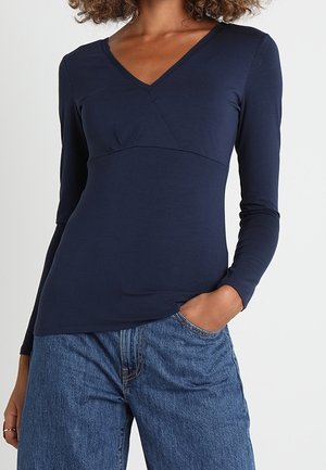 Vrouw draagt een marineblauwe top met lange mouwen en V-hals en losse blauwe spijkerbroek, met één hand in haar zak.