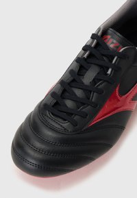 Zapatilla deportiva de cuero negro con acentos rojos, superficie texturizada y detalles cosidos. Presenta cordones negros y un cuello acolchado.