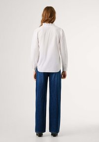 Persona con capelli lunghi che indossa una camicia bianca a maniche lunghe e jeans blu a gamba larga, in piedi con la schiena rivolta, davanti a uno sfondo semplice.