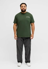 Camiseta de algodón de color verde oliva con mangas cortas y cuello redondo, con un logotipo de texto blanco en el área izquierda del pecho; combinada con pantalones de pana grises.