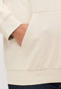 Beige Kapuzenpullover mit einer Kängurutasche, gerippten Bündchen und Saum. Glatte Textur mit dezenten Nähdetails und einer lässigen Passform.