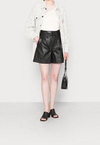 Shorts en cuir noir avec une taille à cordon, associés à une veste blanche texturée et des mules à talons ouverts noires. Petite sac à main noir inclus.