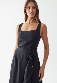 Black linen dress with square neckline, thick straps, cinched waist, and ruffled side detail. Accented with a gold hardware element.
