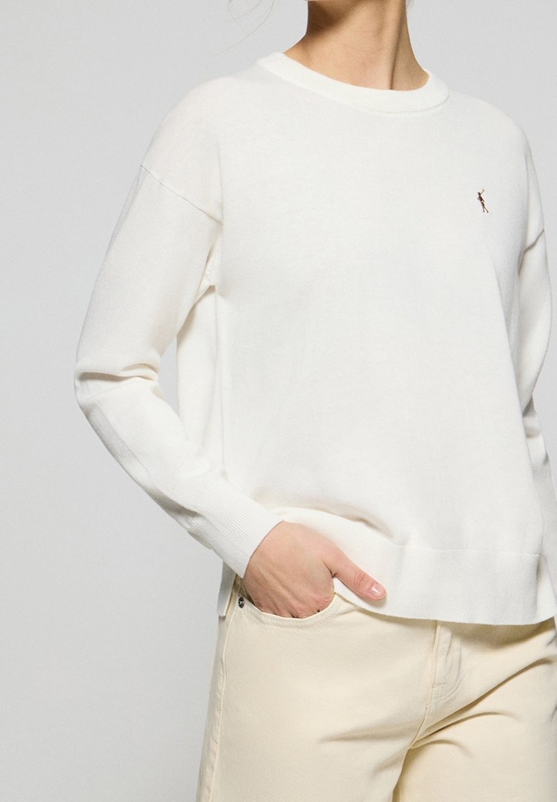 Personne portant un pull blanc à manches longues avec un petit logo brodé sur la poitrine et un pantalon beige clair avec une main dans la poche.