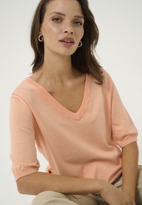 Maglione a maniche corte con scollo a V in morbido tricot pesco. Presenta un colletto a costine e un tessuto texturizzato. Il modello è seduto, con le mani poggiate sulle gambe.