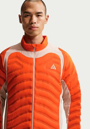 Man wearing bright orange and beige quilted jacket with a front zipper and ACG logo on the chest, standing against a plain background.