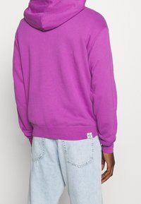 Lila huvtröja med en mjuk textur, med en stor huva, ribbade ärmslut och nederkant. Inkluderar en liten Adidas-logotagg nära nederst.