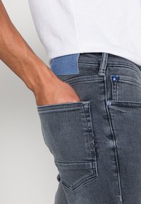 Vaqueros de mezclilla gris oscuro con una etiqueta de cuero azul en la cintura, detalle en el bolsillo delantero y costuras en contraste en el bolsillo trasero.