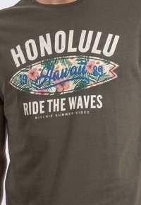 T-shirt olive green à manches courtes avec un graphisme de planche de surf coloré orné de motifs floraux et du texte : "HONOLULU RIDE THE WAVES."