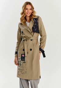 Khaki płaszcz trench z podwójnym zapięciem, czarnymi akcentami i kwiatowymi naszywkami. W talii pasek i dwie boczne kieszenie.