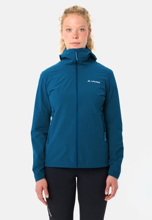 Vrouw met krullend blond haar, gekleed in een blauwe outdoor jas met capuchon en zwarte broek, staand tegen een effen witte achtergrond.