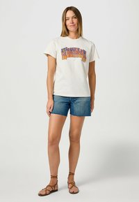 Crème t-shirt met korte mouwen met kleurrijke "Wrangler" graphic. Gepaard met blauwe denim shorts en bruine sandaaltjes met bandjes. Eenvoudige casual outfit.