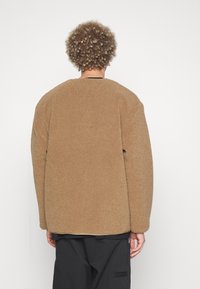Giacca in pile marrone e morbido con una vestibilità ampia e rilassata. Spalle rotonde e maniche lunghe, senza chiusure visibili, indossata sopra pantaloni neri.