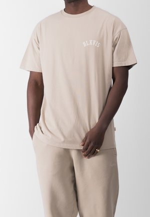 Homme portant un T-shirt à manches courtes beige avec le texte "BLKVIS" et un pantalon beige assorti, une main dans la poche et l'autre reposant sur le pantalon.