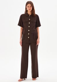 Braun zweiteiliger Anzug mit einem kurzärmeligen, kragenlosen Oberteil mit frontalen Taschen und Knöpfen sowie weiten Hosen. Glatte Textur, minimalistisches Design.
