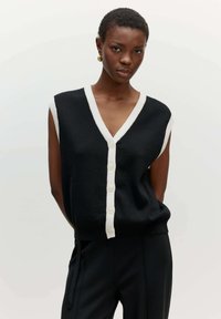 Schwarze ärmellose Strickjacke mit V-Ausschnitt, weißen gerippten Besätzen und sechs großen weißen Knöpfen auf der Vorderseite. Glatte Textur, leichtes Material.
