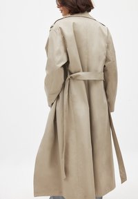 Bežs tranča mētis ar jostu viduklī, garām piedurknēm un vienu apkakli. Izgatavots no gludas auduma, ar taisnu piegriezumu un minimālām detaļām.