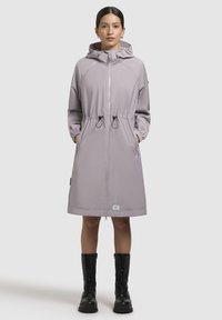 Světle fialový nepromokavý kabát se staženým pasem, kapucí a elastickými manžetami; má přední zip a boční kapsy, v kombinaci s černými botami.