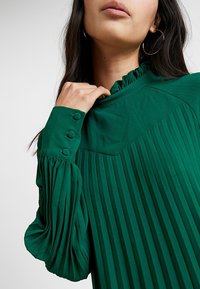 Vihreä pusero, jossa on korkea rypytetty kaulus, pitkät laskostetut hihat ja napin koristeita hihojen päällä. Sileä kangas, jossa on jäykkä muotoilu.