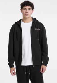 Sort zip-up hoodie lavet af bomuld med hætte og snøre, forlommer og broderet logo på brystet. Parret med en hvid T-shirt.