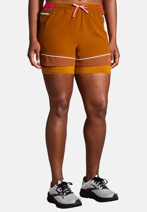 Person iført lagdelte brune og orange atletikshorts med pink snøre og hvide løbesko på en ensfarvet baggrund.