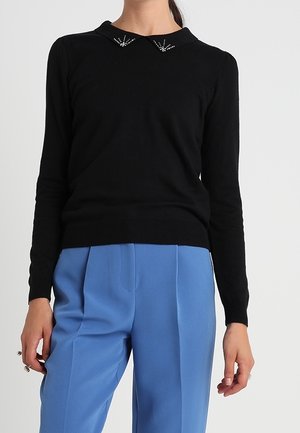 Femme portant un pull noir à manches longues avec des détails décoratifs au col et un pantalon bleu taille haute coupé sur mesure.