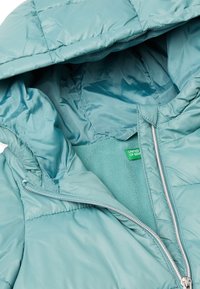 Zelená polstrovaná bunda se zipem, teplou fleecovou podšívkou a kapucí. Obsahuje malou zelenou nálepku s nápisem "UNITED OF BEN."