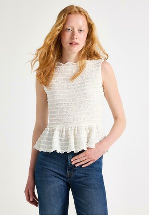 Ung kvinde med langt rødt hår iført en ærmeløs hvid tekstureret top med flæsekant og blå jeans, poserende mod en hvid baggrund.