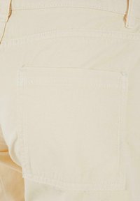 Détails d'un pantalon en coton beige avec une poche arrière et des coutures visibles. Le tissu est lisse et de texture légère.