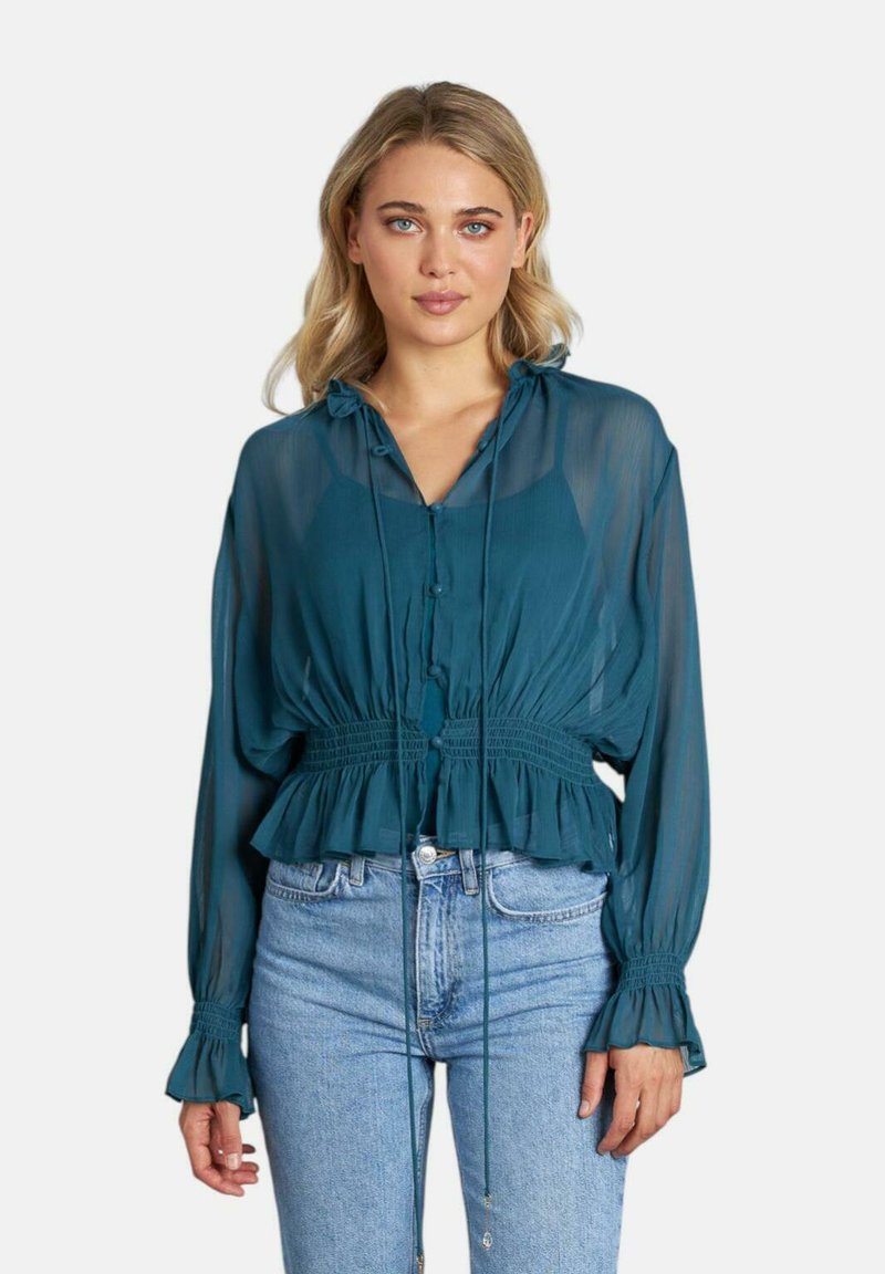 Blusa trasparente teal con maniche lunghe, orlo arricciato e vita regolabile con lacci. Presenta una chiusura con bottoni e polsini accentuati. Indossata con jeans.