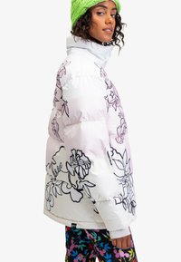 Hvid pufferjakke med pink blomsterdesign, høj krave og sorte kanter. Kombineret med farverige bukser med blomsterdesign og en grøn strikket hue.