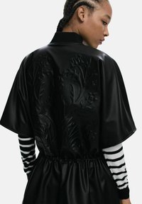 Veste bomber noire avec un motif floral texturé dans le dos, taille élastique froncée, coupe ample et manches longues à rayures contrastantes.
