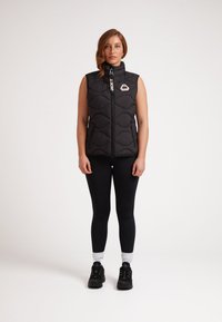 Svart vadderad väst med hög krage och frontdragkedja, med en logotyppatch och en kontrasterande dragkedja. Buren över svarta leggings.