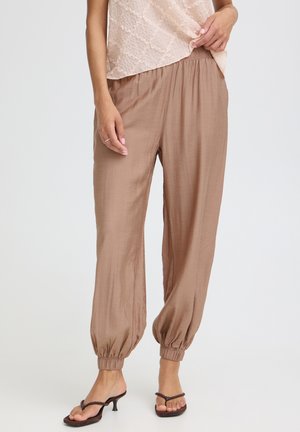 Femme portant un pantalon ample marron clair avec des revers aux chevilles, associé à des sandales à talons marron et un haut texturé rose clair, debout devant un fond blanc.