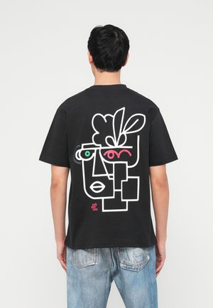 Man iklädd svart t-shirt med färgstarkt abstrakt ansiktsmotiv och ljusblå jeans, sedd bakifrån mot en enfärgad bakgrund.