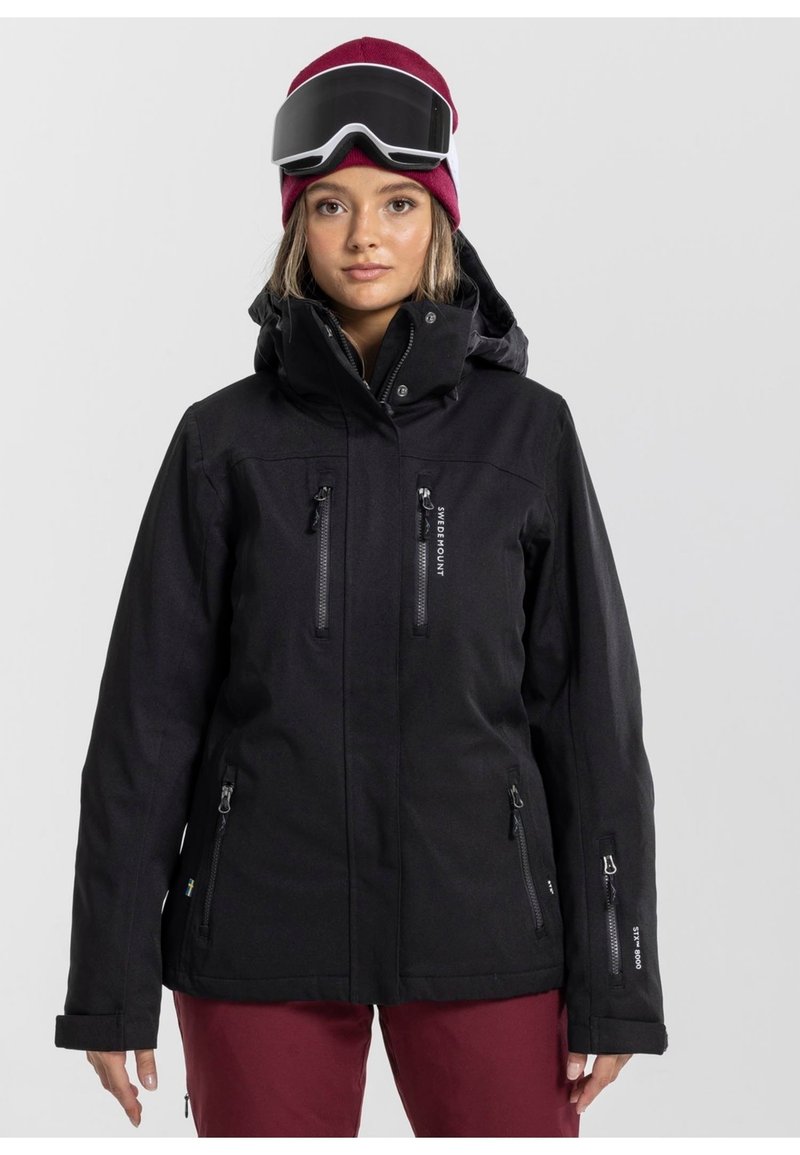Giacca da sci nera impermeabile con cappuccio, tasche con zip e tessuto testurizzato. Indossata con un berretto di maglia bordeaux e occhiali da sci.