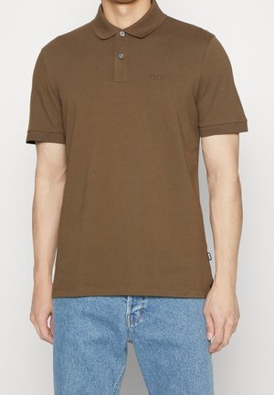 Mies, który ma na sobie brązową koszulę polo z krótkim rękawem z logo "BOSS" na piersi, w połączeniu z jasnoniebieskimi dżinsami, na neutralnym tle.