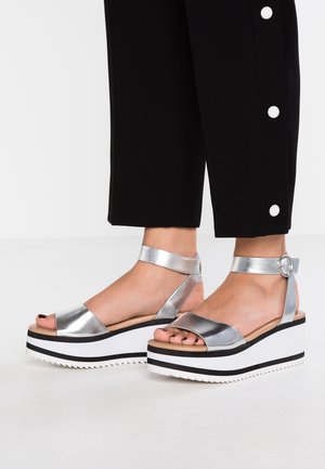 Pieds portant des sandales à plateforme argent métallique avec brides autour de la cheville et semelles épaisses noir et blanc, associées à un pantalon noir avec des boutons blancs.
