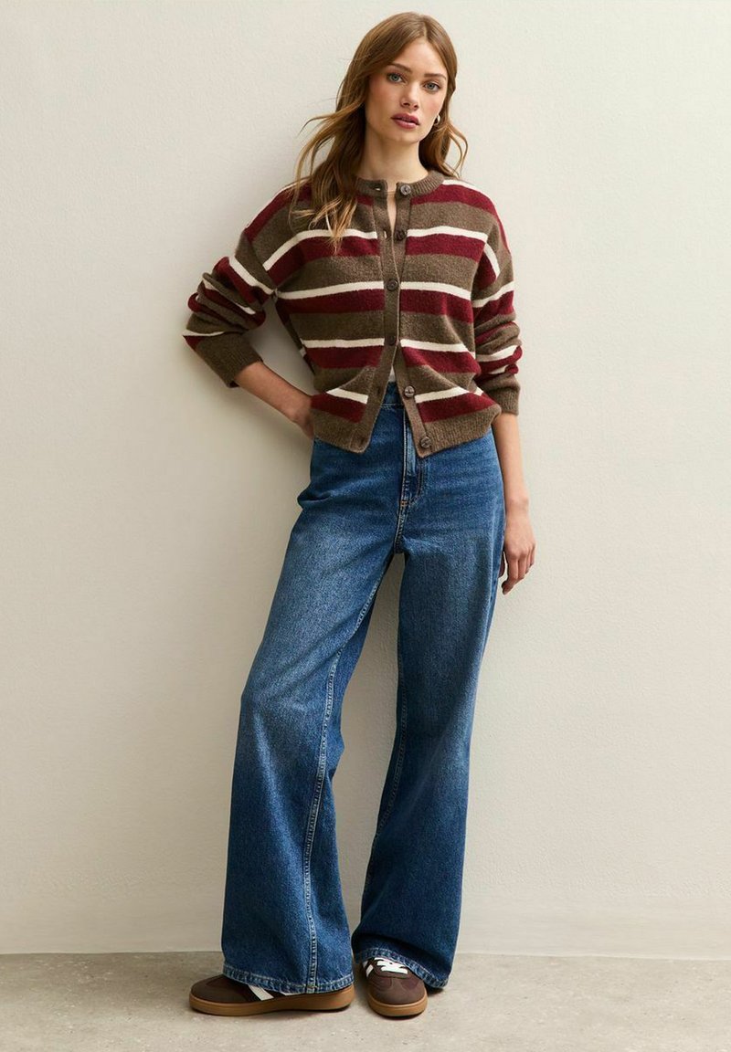 Cardigan rayé en marron, crème et bordeaux avec fermeture à boutons, associé à un jean bleu taille haute et large, ainsi qu'à des baskets marron.