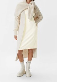 Stickad beige kofta över en ljusgul klänning, matchad med krämiga ankelstövlar. Mjuka texturer, ribbade detaljer och en avslappnad siluett.