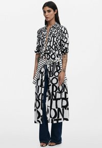 Schwarz-weißes Hemdkleid mit auffälliger, grafischer Schrift, durchgehender Knopfleiste, tailliertem Gürtel und gestreiften Akzenten an Ärmeln und Gürtel.