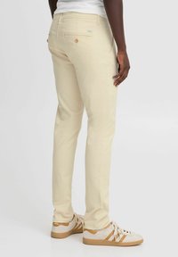 Svijetlosmeđi chinos hlače slim fit kroja, mekane teksture, s stražnjim džepovima sa gumbima. Uparene s bijelim tenisicama s tamnosmešnim detaljima.