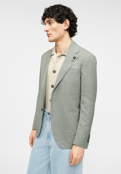Ανοιχτό πράσινο σακάκι σε υφασμάτινη υφή, με κούμπωμα με ένα κουμπί, πέτα με χαραγή και τσέπη στο στήθος, συνδυασμένο με μπεζ πλεκτό πουκάμισο.