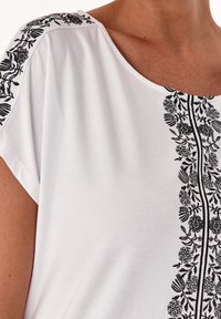 Top blanco de manga corta con bordado floral negro en los hombros y una línea bordada vertical en el frente, textura suave.